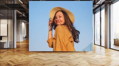 Portrait image of a young woman with hat, hair blows in the wind, glass terrace with blue sky background Wall mural