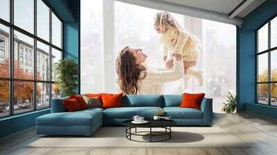 A beautiful mother is playing with happy daughter at the panoramic window in the knitted sweater near the Christmas tree. A young woman hugs little girl, smiling. New year festive card. copy space Wall mural