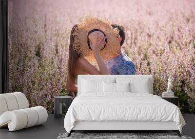 A happy couple in love hid behind a straw hat, kissing at sunset near a blooming sage field. A young man passionately embraces, holds the hands of a beautiful girl. Lovers weekend travel. Copy space Wall mural