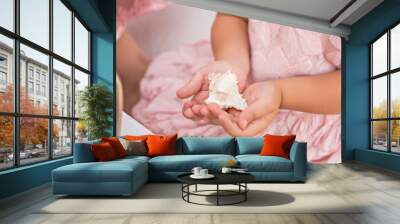 A little cute blonde girl in a gentle pink dress holds a seashell in her hands by the sea. A beautiful baby girl plays with sea treasures. Decoration of celebrations, photo zones in elegant marine sty Wall mural