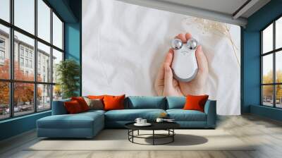 Microcurrent concept. Face massager with micro currents for home use and skincare on white bed. Female hands holds device for anti-aging and face lifting. Top view or flat lay, copy space for text. Wall mural