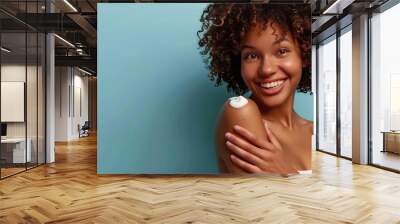 A cheerful young woman with curly hair applying lotion to her shoulder Wall mural