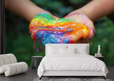 Two hands holding vibrant, multicolored slime dripping and stretching, creating an abstract, playful visual against a blurred green background. Wall mural