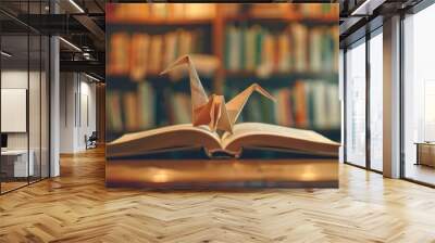 Origami paper crane perched on an open book in a cozy library with a blurred background of bookshelves, symbolizing knowledge and creativity. Wall mural