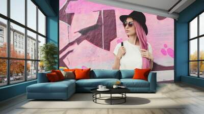 Beautiful stylish young hipster woman with long pink hair, hat and sunglasses on the street. Wall mural