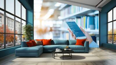 A person using a smartphone in a library, surrounded by shelves of books, showcasing modern technology in an educational environment. Wall mural