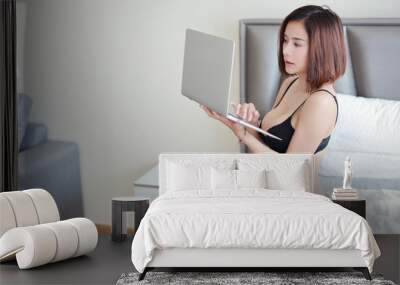 side view of young beautiful and sexy asian woman in black lingerie using laptop computer while sitting on bed in bedroom with relaxation. (internet of thing and life style concept) Wall mural