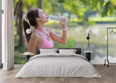 Side view portrait of happy and relax young asian sportswoman take a break after morning workout. Beautiful athlete woman drinking water from bottle after morning run in the park near lake and trees. Wall mural