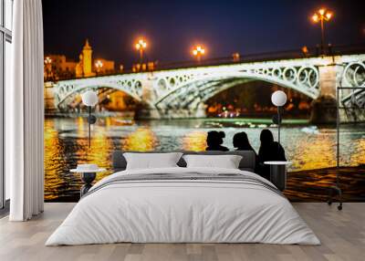 Friends Sitting By Illuminated Bridge In Seville At Night Wall mural