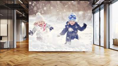 Two young girls, one in a white winter coat and one in a blue winter coat, run through snow playfully. Wall mural