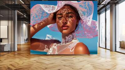 A beautiful young woman in a lace hat poses elegantly under the sun, with delicate shadows casting across her face. A serene and sophisticated summer fashion moment by the sea. Wall mural