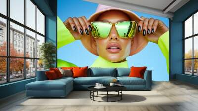 Fashion-forward woman wearing gold reflective sunglasses and a neon green top. Bold and trendy look against a bright blue sky. Wall mural