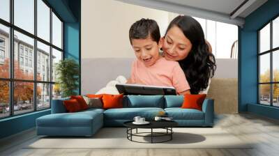 Hispanic mom and her son sitting on the couch at home using a digital tablet while having fun - mom and son watching or playing on the digital tablet - family quality time - single mother Wall mural