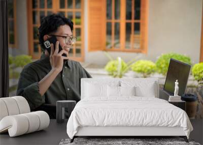 Outdoor shot of young Asian man working on his laptop while talking on mobile phone. Wall mural