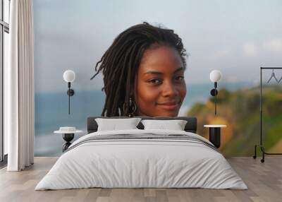 Smiling woman with long dreadlocks wearing earrings and a patterned dress, standing by a scenic coastal landscape, exuding tranquility and natural beauty Wall mural