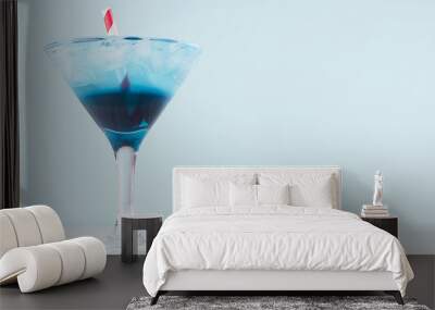 Fresh blue alcohol cocktail with ice cubes, red straw in misted wineglass on green color background, white wood board. Wall mural