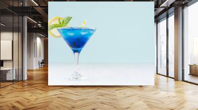 Glamour tropical fruit blue cocktail with curacao, ice cubes, lemon slice, yellow straw, mint in misted martini glass in modern green interior on white wood table. Wall mural