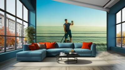 Man in shorts and sunglasses on a paddleboard with a paddle in the sea against the background Wall mural