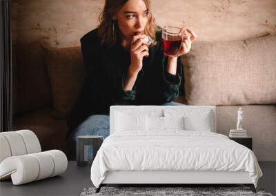 Young thoughtful woman drinking tea and eating chocolate while sitting on sofa at home Wall mural