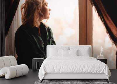 Young thoughtful woman drinking tea and looking through window while sitting on windowsill at home Wall mural