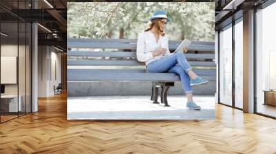 Morning bench / Happy young student with a tablet and a disposable coffee cup sitting on the bench and reading in a summer park. Wall mural