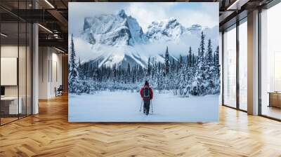 A lone adventurer in winter gear, trudging through a snowy landscape, with a backdrop of snow-capped mountains and pine trees Wall mural