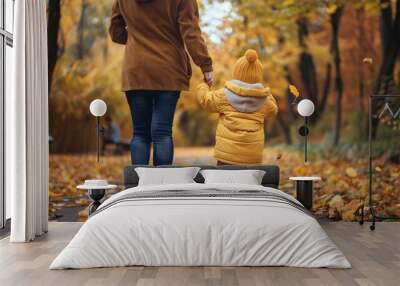 A mother holding a child’s hand as they walk down a park path lined with autumn leaves, both seen from behind Wall mural