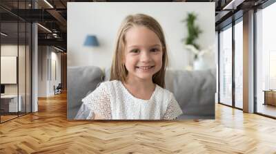 Adorable little girl looking at camera sitting on couch at home, smiling  preschool pretty child with beautiful happy face posing alone on sofa, cute positive cheerful kid headshot portrait Wall mural