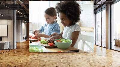 African and Caucasian little girls best friends cooking together in modern kitchen. Multiracial cousins hold knives cutting vegetables on wooden board prepare healthy salad making surprise for parents Wall mural