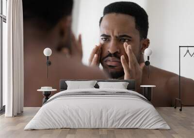 Anxious young african American man look in mirror in bathroom concerned about face wrinkles, frustrated unhappy biracial male worried about acne, unhealthy facial skin, skincare concept Wall mural