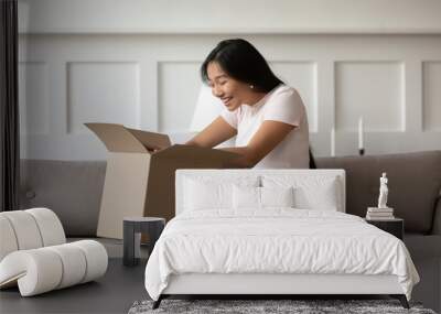 Asian woman sitting on couch in living room opens received delivered parcel feels happy. Satisfied client shopper buy qualified goods in internet unbox package, quick trusted delivery service concept Wall mural