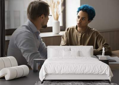 Attractive blue-haired African employee lead talk to male colleague seated at desk in office, looks serious while consider issue solution, exchanging opinions, ideas and creative thoughts at workplace Wall mural