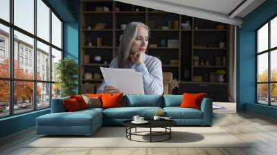 Attractive brooding aged businesswoman sit at workplace desk holds papers feels exhausted distracted from paperwork, staring aside looks serious and thoughtful. Overload, overwork, concerns concept Wall mural