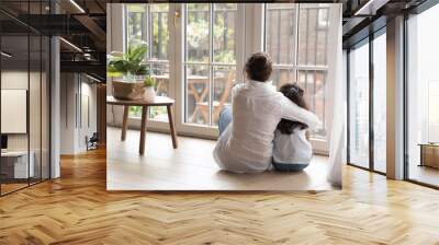 Back view of black haired young mom and little daughter girl sitting on floor at home, hugging with love, care, looking at panoramic window, cozy balcony. Motherhood, real estate concept Wall mural