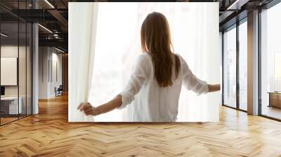 Back view of millennial woman open white curtains in modern light apartment, happy female look out of window enjoying beautiful city view, admiring skyscrapers or watching people during sunny morning Wall mural