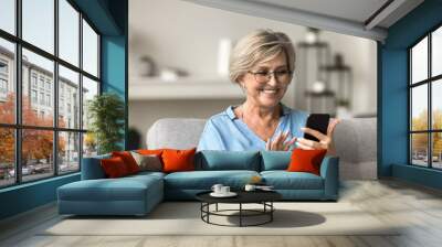 Beautiful older female wear glasses enjoy remote chat, on-line communication with family using smartphone, sit on sofa in living room, read news, watch internet content, spend carefree leisure at home Wall mural