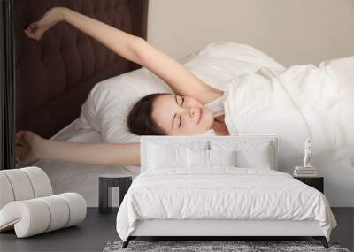 Beautiful woman stretching with pleasure on white sheets. Teenage girl feeling positive waking up early in the morning after good night sleep. Healthy sleep, good beginning of wonderful day concept Wall mural