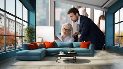 Business people using computer, millennial male employee in formal suit helping explaining to middle aged mature female colleague learn understand corporate program. Mentoring and assistance concept Wall mural