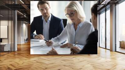 Businessmen sitting at desk headed by middle aged serious concentrated female in eyeglasses checking agreement document before signing it. Financial director ready affirm official paper with signature Wall mural