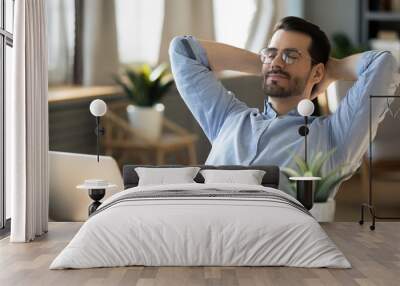 Calm millennial man in glasses sit relax at home office workplace take nap or daydream. Happy relaxed Caucasian young male rest in chair distracted from computer work, relieve negative emotions. Wall mural