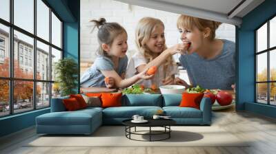 Caring daughter feeding mother teaching little children standing at table in kitchen. Happy mom with girls sisters chopping vegetables lettuce on wooden cutting board, preparing dinner. Wall mural