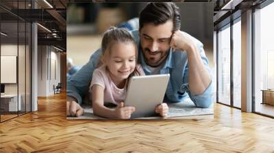 Caring young father cuddling small 6 years old daughter, lying together on floor using digital tablet at home. Happy little cute kid girl relaxing with dad, watching funny cartoons on gadget. Wall mural