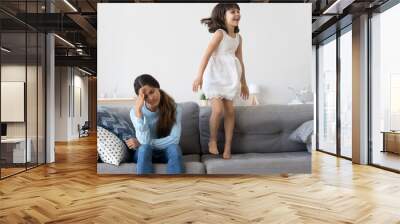 Cheerful active daughter have fun jumping on sofa. Tired single frustrated woman holds her head suffering from headache, child annoy her. Feeling unwell or depression, education difficulties concept Wall mural