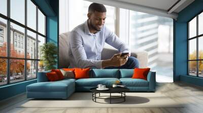 Cheerful attractive young Black man enjoying Internet communication at home, holding mobile phone, using online application, sitting on sofa in city apartment, looking at screen, smiling Wall mural