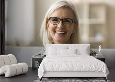 Cheerful beautiful blonde mature woman in trendy eyeglasses and casual shirt looking at camera, smiling, showing white teeth. Happy senior freelance business lady head shot video call portrait Wall mural