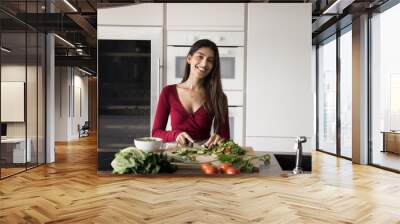 Cheerful beautiful young Indian chef woman preparing healthy lunch, meal from organic food, chopping green vegetables on board, cooking dinner in home kitchen, looking at camera, smiling for portrait Wall mural