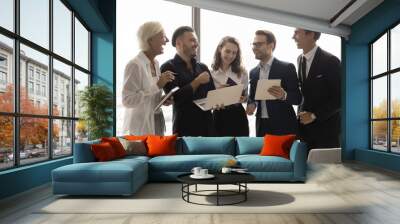 Cheerful business colleagues working on project tasks together, using laptop, tablet for cooperation, discussing online presentation, laughing, standing by large office window, enjoying teamwork Wall mural