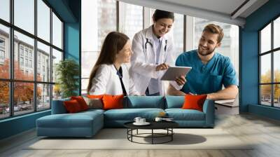 Cheerful diverse clinic medical colleagues in uniforms using tablet computer at workplace together, talking, laughing, discussing healthcare online service, enjoying modern Internet technology Wall mural