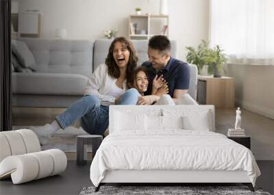 Cheerful excited parents tickling beloved kid girl, playing with child on heating floor in modern home interior, sitting on carpet, laughing, shouting for joy, enjoying family leisure Wall mural