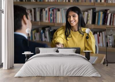 Cheerful Indian student woman talking to male professor in college library, giving handshake to tutor, teacher, thanking for help in study research, sitting at table with bookshelves behind Wall mural